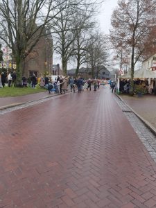 Nasse gepflasterte Straße mit Menschen, die sich links bei einem geschmückten Weihnachtsbaum vor einer Kirche und rechts an einem Verkaufszelt versammeln.