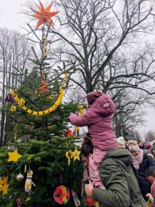 Ein Kind in rosa Schneeanzug sitzt auf den Schultern eines Erwachsenen und hängt selbstgebastelten Schmuck an einen geschmückten Weihnachtsbaum im Freien.