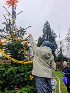 Ein Kind in Winterkleidung steht auf einer kleinen Leiter und hängt eine gelbe Papierkette an einen mit Stern geschmückten Weihnachtsbaum im Freien.