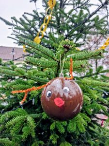 Nahaufnahme einer braunen Weihnachtskugel mit aufgemaltem Gesicht und Pfeifenputzer-Geweih, die an einem geschmückten Tannenbaum hängt.