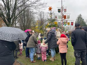 Viele Menschen mit Kindern und Regenschirmen versammeln sich im Nieselregen um einen geschmückten Weihnachtsbaum neben einem Maibaum mit bunten Vereinswappen.
