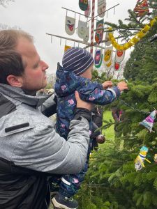 Ein Mann hält ein Kleinkind im bunten Schneeanzug, das an einem geschmückten Weihnachtsbaum im Freien schmückt.