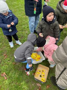 Mehrere kleine Kinder in Winterkleidung greifen nach buntem Weihnachtsschmuck aus einer Kiste auf dem Rasen.