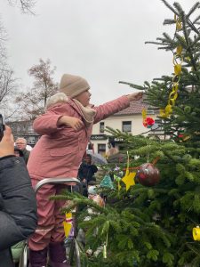 Ein Kind in rosa Schneeanzug steht auf einer Leiter und hängt Weihnachtsdeko an einen geschmückten Tannenbaum.