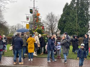 Viele Menschen mit Kindern versammeln sich bei grauem Wetter um einen geschmückten Weihnachtsbaum und einen Maibaum auf einer Wiese.