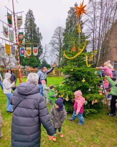 Kinder und Erwachsene schmücken gemeinsam einen Weihnachtsbaum auf einer Wiese neben einem mit bunten Vereinswappen behängten Maibaum.