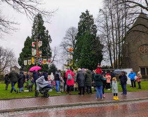 Eine große Gruppe von Menschen mit Kindern versammelt sich bei Nieselregen um einen geschmückten Weihnachtsbaum und einen Maibaum vor einer Kirche.