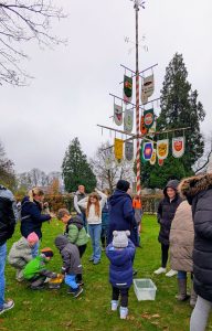 Kinder und Erwachsene stehen und spielen auf einer Wiese rund um einen mit bunten Vereinswappen geschmückten Maibaum.