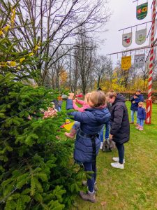 Kinder und Erwachsene schmücken gemeinsam einen Weihnachtsbaum im Freien neben einem mit Wappen geschmückten Maibaum.